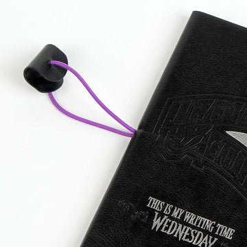 Schwarzer Schein Notizblock Wednesday mit lila Gummiband und geprägtem Schriftzug.