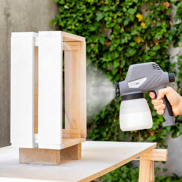 Graue elektrische Farbspritzpistole Spraint+ beim Anbringen von weißer Farbe auf einen Holzbehälter.