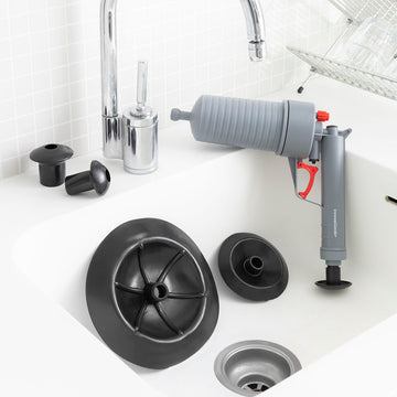 Graue Druckluftpistole zur Abflussreinigung mit schwarzen Adaptern im Waschbecken.